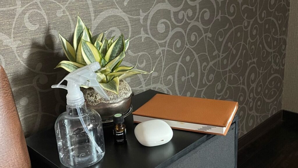 ベッドサイドのサイドテーブルに無印アロマウォーター用のスプレーボトルと精油、本、観葉植物を置いたリラックスできる寝室の一角