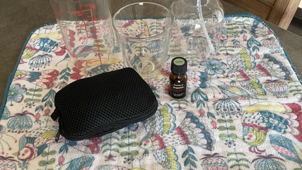 エタノールなしでアロマスプレーを作るために、空ボトルと精油瓶、水を入れたコップや計量カップをマットの上に並べた手作り準備シーン